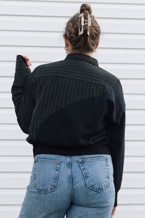 A woman viewed from behind wearing a black color-blocked long-sleeve top with a ribbed yoke and blue jeans, standing against a white horizontal siding wall.