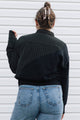 A woman viewed from behind wearing a black color-blocked long-sleeve top with a ribbed yoke and blue jeans, standing against a white horizontal siding wall.