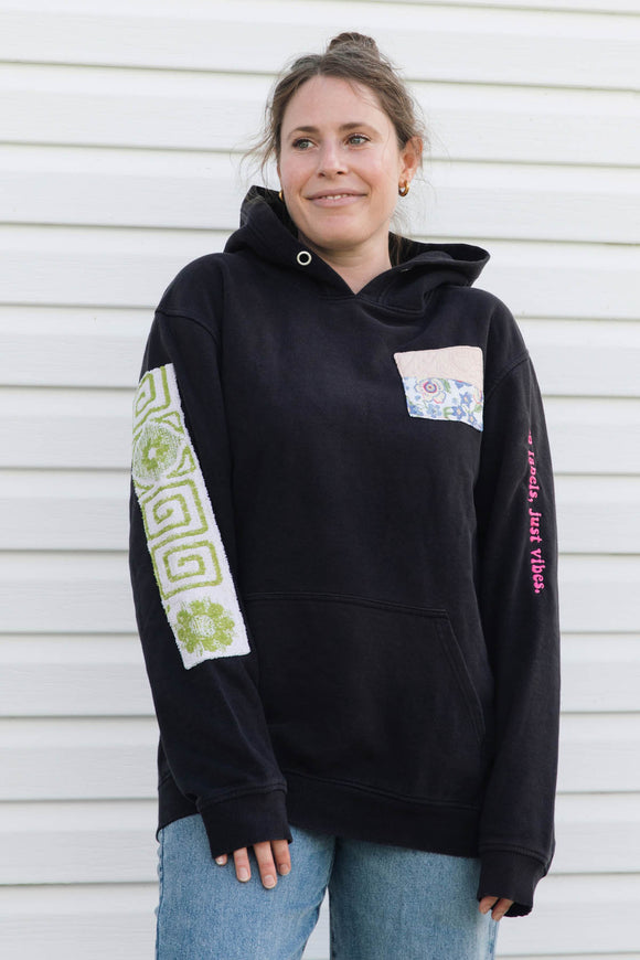 A woman wearing a black pullover hoodie with decorative patchwork details on the sleeves and chest pocket, paired with blue jeans, standing against a white siding background.