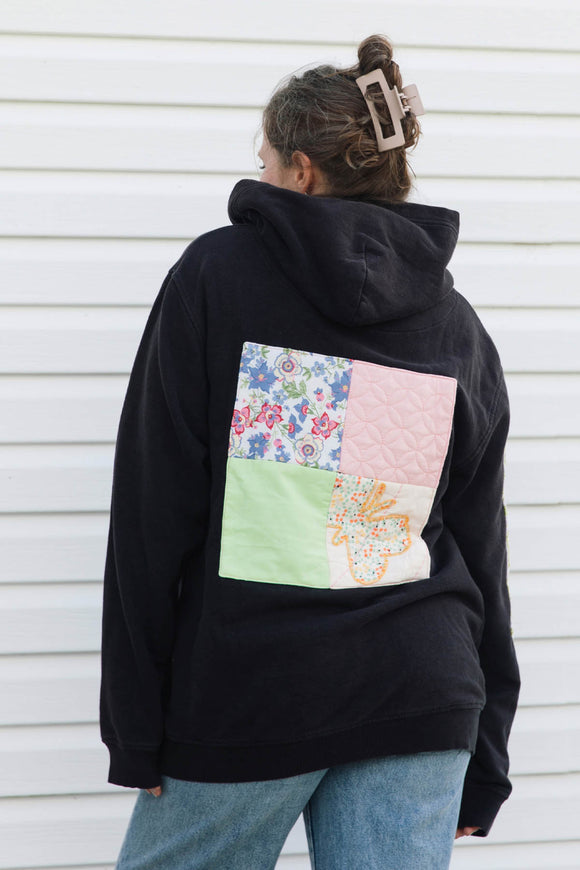 A person viewed from behind wearing a black hoodie with a colorful patchwork quilt design on the back, paired with blue jeans, standing against a white siding wall.