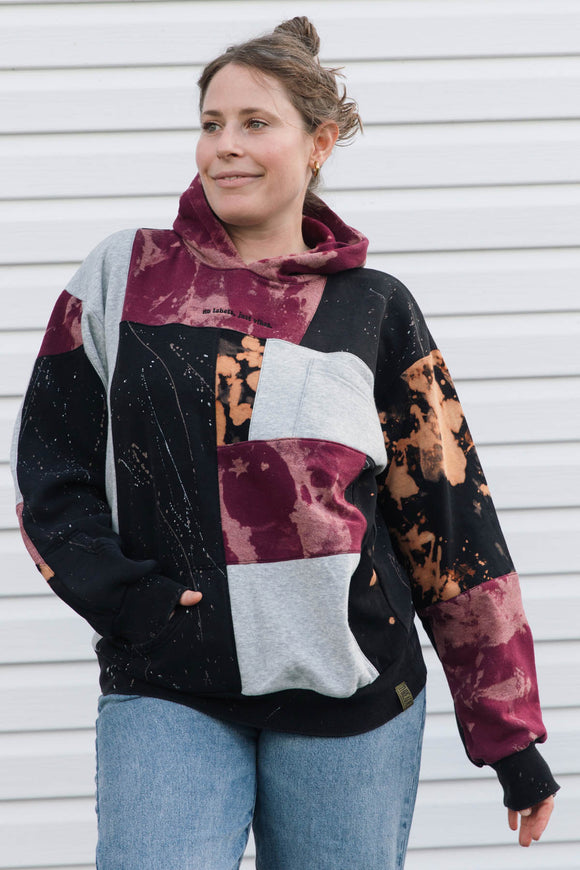 A woman wearing a patchwork hoodie with color-blocked panels of black, heather gray, burgundy, and orange-brown tie-dye patterns, paired with blue jeans, standing against a white siding background.