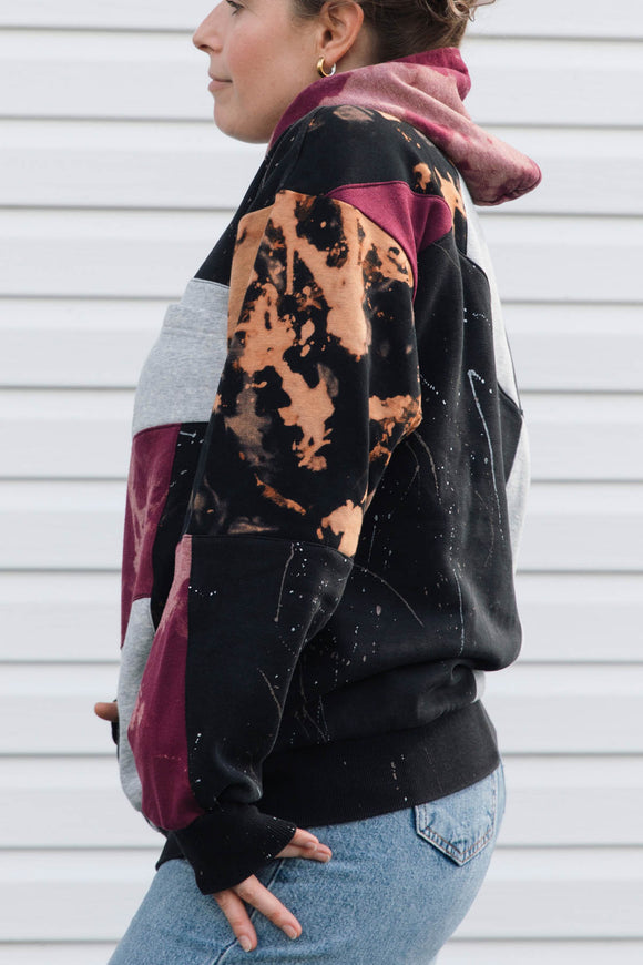 A woman shown from the side wearing a color-blocked hoodie with a black base, maroon panels, a light gray panel, and a tan-and-black tie-dye pattern on the upper sleeve, paired with blue jeans in front of a white siding background.