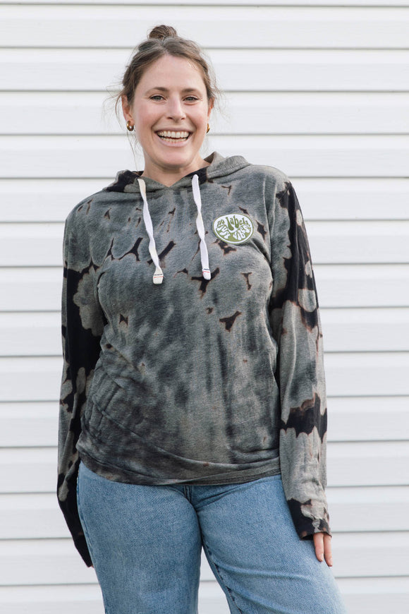A woman smiling while wearing a dark gray and black acid-wash tie-dye hoodie with white drawstrings and a circular green and white logo patch on the left chest, paired with blue jeans, standing in front of a white siding wall.