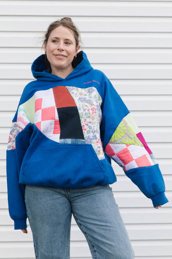A woman wearing a blue hoodie with a colorful patchwork quilt design on the chest and sleeves, paired with blue jeans, standing in front of a white siding wall.