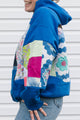 A person wearing a bright blue hoodie with colorful patchwork quilted sleeves and back panels, paired with blue jeans, standing against a white siding background.