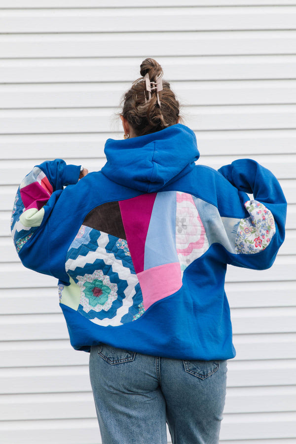 A person viewed from behind wearing a bright blue hoodie with colorful patchwork quilted details on the back and sleeves, paired with blue jeans, standing against a white horizontal siding wall.