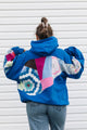 A person viewed from behind wearing a bright blue hoodie with colorful patchwork quilted details on the back and sleeves, paired with blue jeans, standing against a white horizontal siding wall.