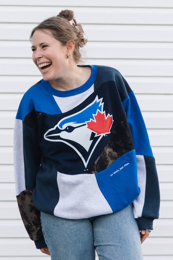 A woman wearing a Toronto Blue Jays colorblock sweatshirt, laughing against a white siding background.