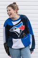 A woman wearing a Toronto Blue Jays colorblock sweatshirt, laughing against a white siding background.
