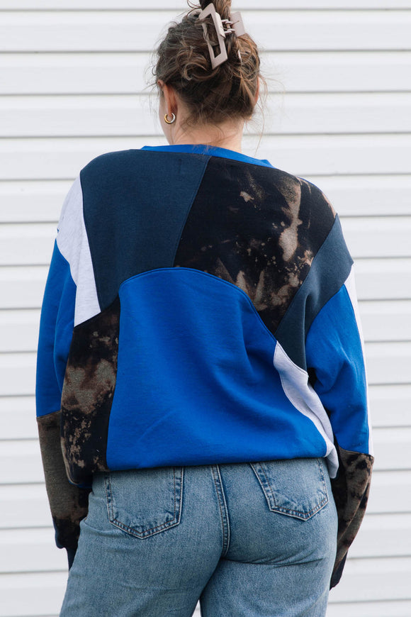 A woman viewed from behind wearing a color-block crewneck sweatshirt with blue, black, and white panels, paired with light-wash blue jeans against a white siding background.