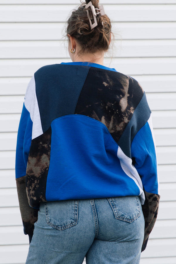A woman viewed from behind wearing a color-block crewneck sweatshirt with blue, black, and white panels, paired with light-wash blue jeans against a white siding background.