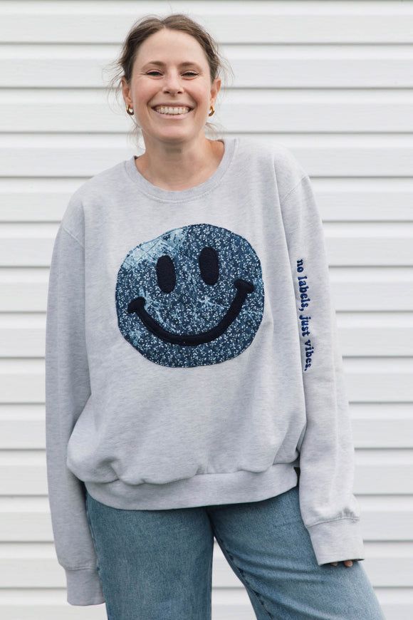 A woman smiling while wearing a light gray crewneck sweatshirt with a large blue glittery smiley face on the front and blue jeans, standing against a white siding background.