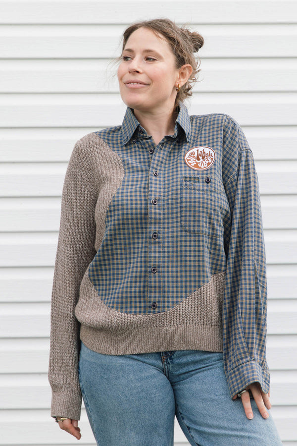 A woman wearing a long-sleeve button-up shirt with a blue and brown gingham body and ribbed taupe knit sleeves and hem, paired with blue jeans, standing against a white siding background.