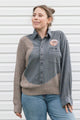 A woman wearing a long-sleeve button-up shirt with a blue and brown gingham body and ribbed taupe knit sleeves and hem, paired with blue jeans, standing against a white siding background.