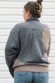 A woman viewed from behind wearing a patchwork button-up shirt with a blue and beige gingham body and ribbed beige sleeves, collar, and hem, paired with blue jeans against a white siding background.