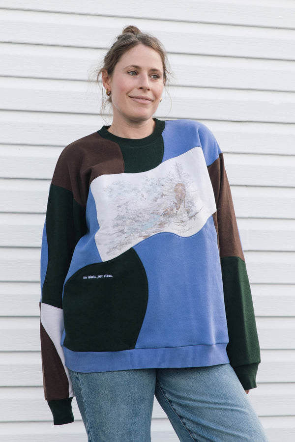 A woman wearing a color-blocked crewneck sweatshirt with a large abstract art print on the front, paired with blue jeans, standing against a white horizontal siding wall.
