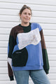 A woman wearing a color-blocked crewneck sweatshirt with a large abstract art print on the front, paired with blue jeans, standing against a white horizontal siding wall.