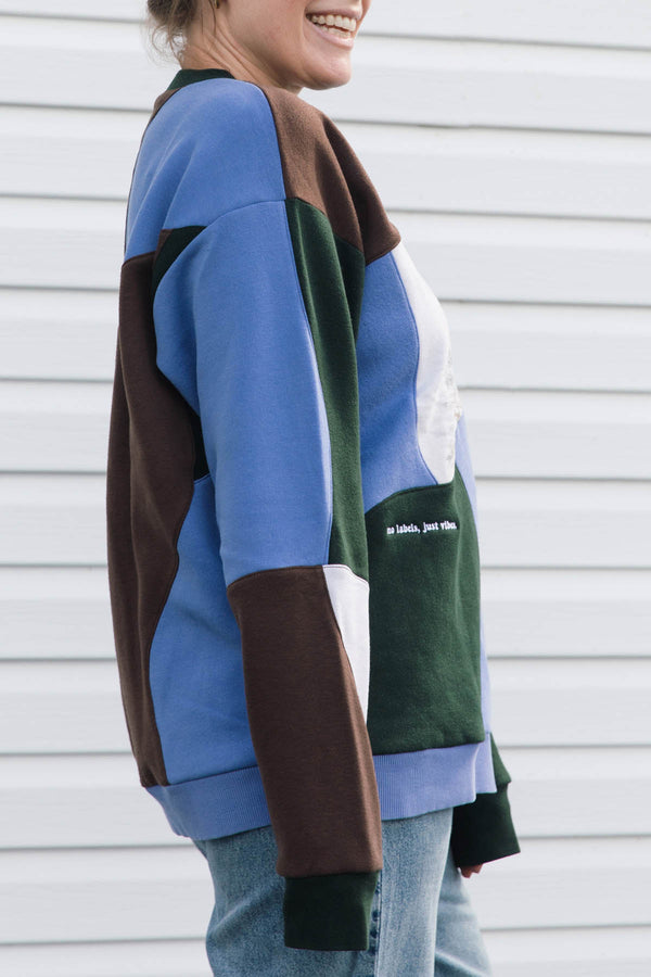 A person wearing a color-block knit sweater with panels of blue, brown, dark green, and white, paired with light blue jeans, standing against a white siding wall.