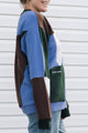 A person wearing a color-block knit sweater with panels of blue, brown, dark green, and white, paired with light blue jeans, standing against a white siding wall.