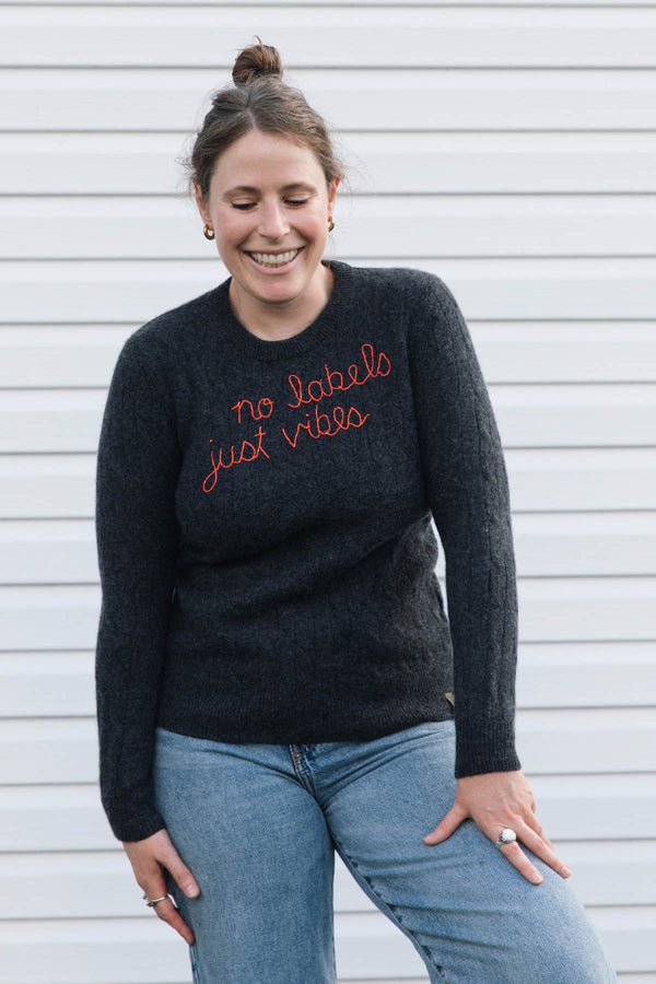 A woman smiling while wearing a dark charcoal gray knit sweater with orange cursive text that reads 'no labels just vibes' and light blue jeans, standing against a white siding background.