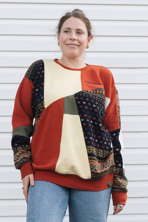 A woman wearing a retro-style color-blocked crewneck sweatshirt with geometric panels of rusty orange, yellow, olive green, and dark patterned fabric, paired with blue jeans, standing in front of a white siding wall.