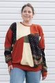 A woman wearing a retro-style color-blocked crewneck sweatshirt with geometric panels of rusty orange, yellow, olive green, and dark patterned fabric, paired with blue jeans, standing in front of a white siding wall.