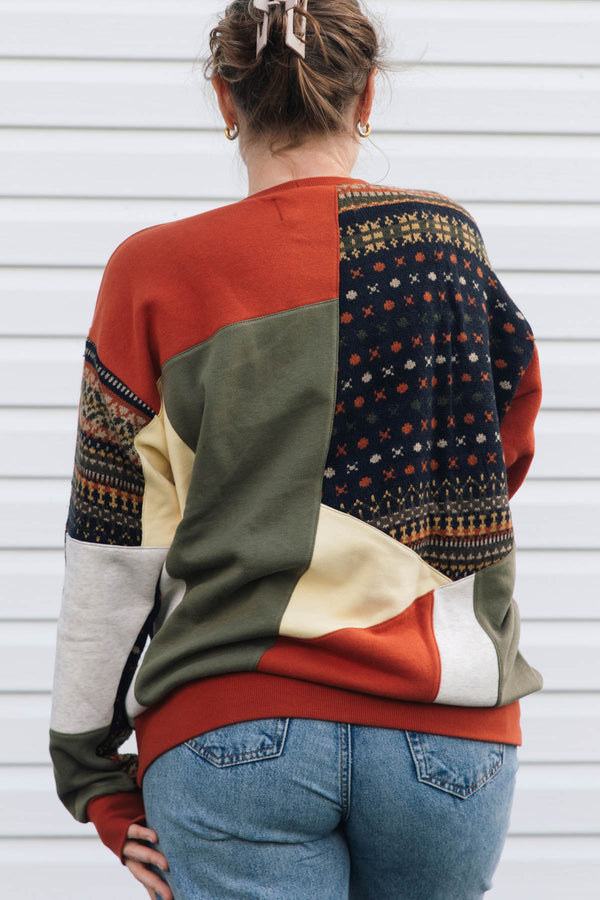 A woman viewed from behind wearing a color-block knit sweater with geometric panels in rust orange, olive green, cream, and a dark navy patterned section, paired with blue jeans against a white siding background.
