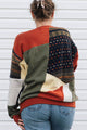 A woman viewed from behind wearing a color-block knit sweater with geometric panels in rust orange, olive green, cream, and a dark navy patterned section, paired with blue jeans against a white siding background.