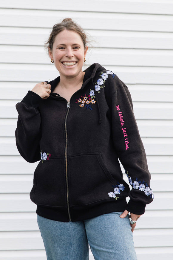 A woman smiling while wearing a black zip-up hoodie with colorful floral embroidery and pink text on the sleeve, paired with blue jeans, standing against a white siding background.