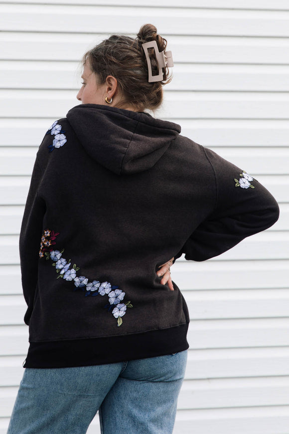 A woman wearing a black hoodie with embroidered blue and white flowers on the back and shoulders, paired with blue jeans, standing against a white siding background.