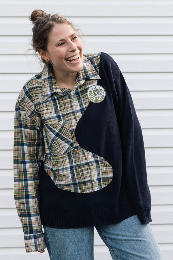 A woman wearing a unique patchwork sweater made from a navy knit and a beige plaid shirt, standing in front of a white siding background.