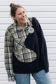 A woman wearing a unique patchwork sweater made from a navy knit and a beige plaid shirt, standing in front of a white siding background.