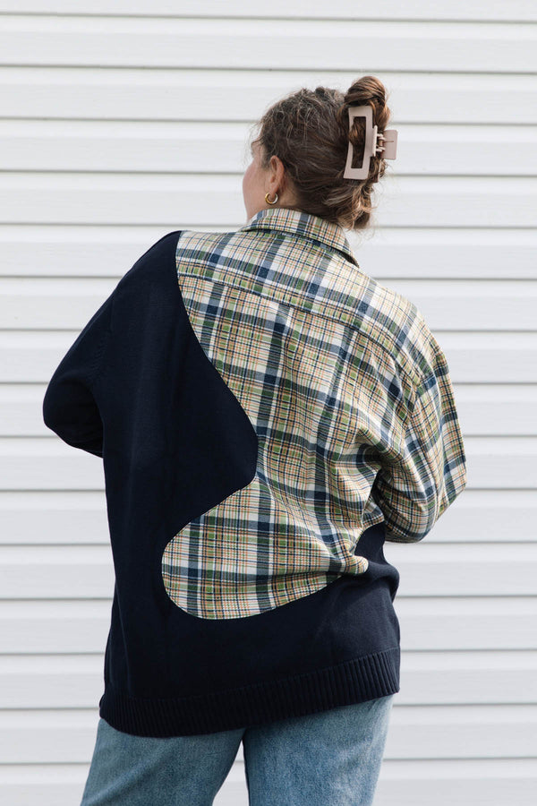 A person shown from the back wearing a color-block knit sweater over a plaid button-up shirt and blue jeans, standing against a white horizontal siding wall.