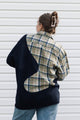 A person shown from the back wearing a color-block knit sweater over a plaid button-up shirt and blue jeans, standing against a white horizontal siding wall.