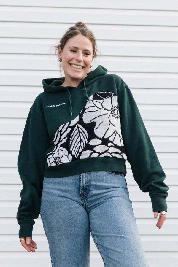 A woman wearing a dark green cropped hoodie with a large black and white floral pattern on the front, paired with light blue jeans, standing against a white horizontal siding background.