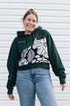 A woman wearing a dark green cropped hoodie with a large black and white floral pattern on the front, paired with light blue jeans, standing against a white horizontal siding background.