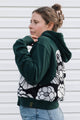 A woman wearing a dark green hoodie with a black and white floral knit pattern on the back, paired with blue jeans, standing against a white siding background.