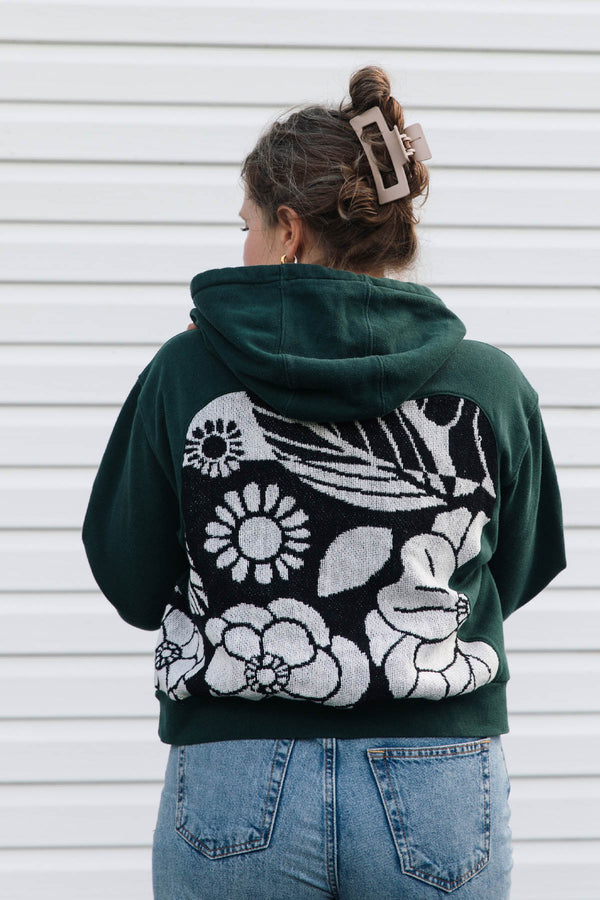 A woman viewed from behind wearing a dark green cropped hoodie with a large black and white floral knit pattern across the back, paired with blue jeans against a white siding background.