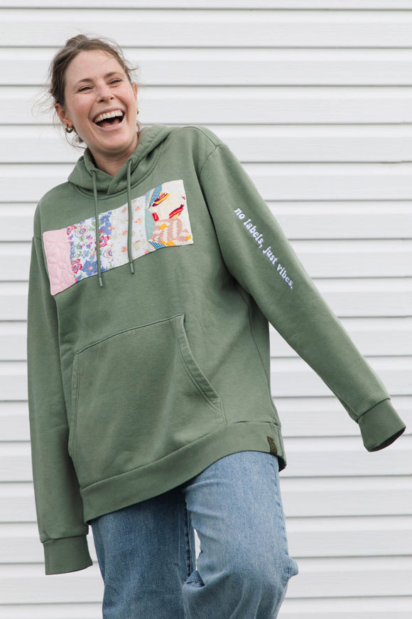 A woman laughing while wearing an oversized sage green hoodie with a colorful floral patchwork graphic on the chest and white script text on the sleeve, paired with blue jeans, set against a white siding background.