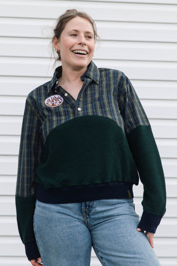 A woman smiling while wearing a color-blocked button-up shirt with a green and navy plaid upper section, a solid dark green middle panel, and dark green sleeves, paired with light blue jeans, set against a white siding background.