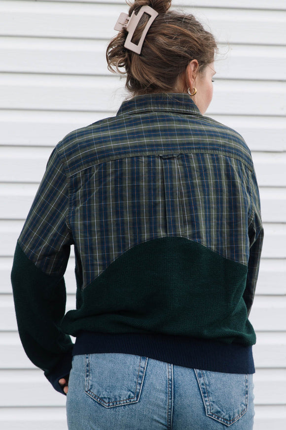 A woman viewed from behind wearing a color-block plaid shirt jacket with navy, green, and yellow plaid on the upper back and shoulders, solid dark green sleeves and lower back, and a navy ribbed hem, paired with blue jeans against a white siding background.