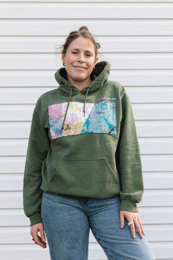 A woman wearing an olive green pullover hoodie with a colorful rectangular graphic print on the chest, paired with blue jeans, standing against a white horizontal siding wall.