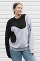 A woman wearing a color-blocked knit sweater with a black yoke, black abstract patches, and a light gray body with blue text on the sleeve, paired with blue jeans, standing against a white siding background.
