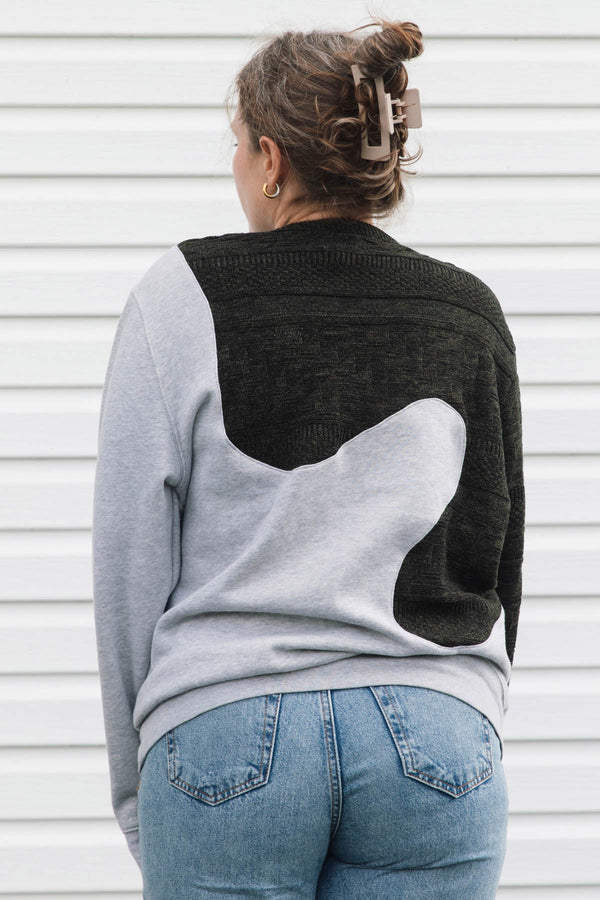 A woman viewed from behind wearing a color-block knit sweater with light gray sleeves and body, a black yoke, and a large white abstract patch across the back, paired with blue jeans against a white siding background.