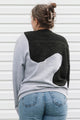 A woman viewed from behind wearing a color-block knit sweater with light gray sleeves and body, a black yoke, and a large white abstract patch across the back, paired with blue jeans against a white siding background.
