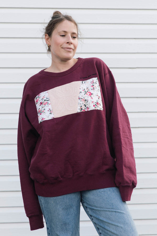 A woman wearing a burgundy crewneck sweatshirt with a beige and floral patchwork chest design, paired with blue jeans, standing against a white siding wall.