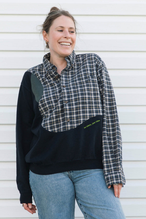 A woman wearing a color-blocked button-up shirt sweatshirt with a plaid pattern on the chest and sleeves, solid black on the sleeves and lower body, and light blue jeans, standing against a white siding background.