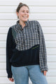 A woman wearing a color-blocked button-up shirt sweatshirt with a plaid pattern on the chest and sleeves, solid black on the sleeves and lower body, and light blue jeans, standing against a white siding background.