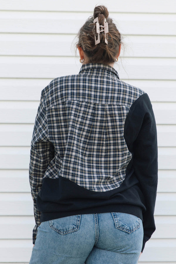 A woman viewed from behind wearing a color-blocked plaid and solid long-sleeve shirt, paired with blue jeans and a beige claw clip, standing against a white siding wall.