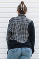 A woman viewed from behind wearing a color-blocked plaid and solid long-sleeve shirt, paired with blue jeans and a beige claw clip, standing against a white siding wall.